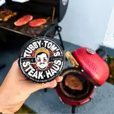 A close-up, first-person perspective shot of a hand holding a tin of Tubby Tom's Steak Haus seasoning. In the background, a BBQ grill with raw steaks and a red Kamado-style grill are visible, establishing a home cooking and outdoor grilling context.