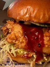 A close-up, macro-style shot of a fried chicken burger, highlighting the texture of the crispy coating and the glossy sauce. The image focuses heavily on the main components of the burger.