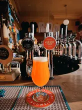 A well-composed, eye-level shot of a glass of beer resting on a bar counter, with the pub's beer taps and interior visible in the soft-focus background. The warm lighting enhances the golden hue of the drink, creating an inviting and authentic pub atmosphere.