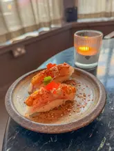 A close-up shot of two pieces of seared nigiri sushi served on a rustic ceramic plate, accompanied by a lit candle in the background. The image captures a dining moment at a Neo-Nikkei restaurant, highlighting the presentation of the sushi with a focus on the textures of the fish and the garnish.