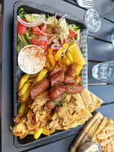 A high-quality, overhead flat-lay photograph of a Greek mixed grill platter served on a tray lined with blue gingham paper. The dish includes grilled chicken, sausages, fries, a fresh salad, tzatziki, and pita bread, presented in a casual, inviting manner.