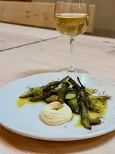 A high-quality, minimalist food photograph featuring a plated vegetable dish alongside a glass of white wine on a light wooden table. The composition is clean and professional, highlighting the fresh ingredients and elegant presentation typical of a farm-to-table dining experience.