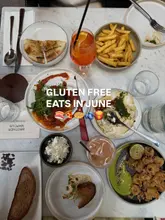 A high-quality, overhead flat-lay photograph showcasing a variety of gluten-free dishes and drinks on a marble table, likely taken at a Mediterranean-style restaurant. The image effectively communicates a 'feast' or 'sharing' dining experience.