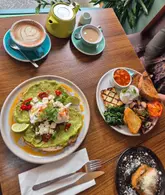 A high-quality, overhead flat-lay photograph of a brunch spread featuring avocado toast, a vegetarian breakfast plate, a latte, and tea. The lighting is soft and natural, highlighting the textures and colours of the food effectively.