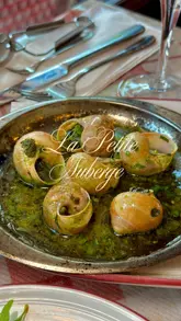 A montage video showcasing a variety of French dishes, cocktails, and the interior/exterior of La Petite Auberge. The video features close-up shots of food, including escargots, grilled octopus, steak, prawns, and a tableside preparation of Crêpe Suzette, set to French-style music.
