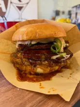 A close-up, eye-level shot of a stacked burger served in a paper wrapper, highlighting the glaze and pork belly topping.