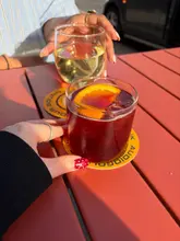 A casual, first-person perspective shot of two people enjoying drinks outdoors at sunset. The image features a red cocktail in the foreground and a glass of white wine in the background, set on a red table with branded coasters.