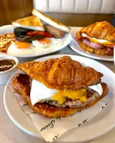 A high-quality, close-up shot of a breakfast croissant sandwich featuring a sausage patty, egg, and melted cheese, served on branded paper. The shallow depth of field keeps the focus sharp on the foreground item while softly blurring the full English breakfast and additional sandwich in the background, creating an inviting and cohesive breakfast scene.