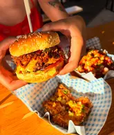 A close-up shot of a person holding a loaded burger, with sides visible in the foreground. The image captures a casual street food dining experience at an outdoor venue.