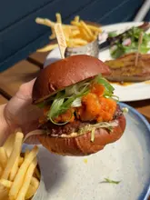 A close-up, handheld shot of a surf and turf burger featuring a beef patty topped with shrimp, held up against a restaurant background.