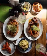 A high-quality, top-down flat lay photograph of a Sunday roast spread at a pub. The image features two large Yorkshire puddings filled with roast meats and vegetables, accompanied by sides like mac and cheese, onion rings, and roasted carrots, along with cocktails. The lighting is natural and warm, highlighting the textures of the food.