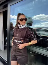 A high-quality, well-composed portrait of a woman posing in a McLaren Racing shirt, likely taken at a hospitality suite during the British Grand Prix at a Hilton venue. The image is sharp, well-lit, and professional, effectively capturing a lifestyle and fashion moment within a premium event setting.