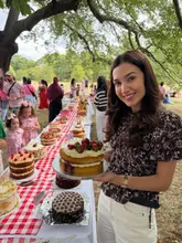 A woman poses with a strawberry shortcake at an outdoor picnic event, surrounded by a long table filled with various cakes. The setting is casual and festive, featuring a red gingham tablecloth and a park-like environment.