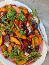 A vibrant, overhead shot of a stone fruit caprese salad featuring sliced peaches, cherries, mozzarella, and arugula, served on a white platter with a serving spoon.