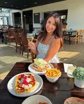 A lifestyle-focused shot featuring a woman smiling and holding a glass of white wine at a restaurant table, with two flatbreads and a bowl of fries in the foreground.