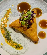 A close-up, top-down shot of a samosa chaat dish served on a textured plate, featuring a samosa topped with microgreens, surrounded by dots of sauce and a smear of yogurt-based dressing.