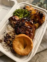A top-down, well-lit shot of a Caribbean-style roast dinner served in a compostable takeaway container, featuring jerk chicken, rice and peas, roasted vegetables, and a Yorkshire pudding.