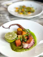A high-quality, professional food photograph showcasing a beautifully plated grilled octopus dish served on a vibrant green puree with crispy potato cubes and a fresh lime wedge, captured in a restaurant setting.