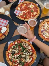 A high-angle, flat-lay shot capturing a group dining experience featuring multiple pizzas and pints of beer on an outdoor table. The image effectively conveys a casual, social atmosphere suitable for friends or groups.