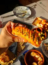 A vibrant, golden-hour close-up shot of a hand holding a piece of tapas, likely a croqueta, topped with a rich sauce and chives. The background features a table filled with various Spanish dishes, creating an inviting and social dining atmosphere.