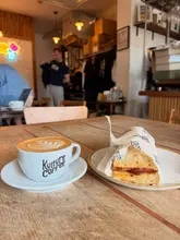 A casual, well-lit shot of a coffee and a bacon sandwich on a wooden table at a cafe, showcasing the venue's branding on the cup and packaging.