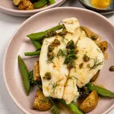 A high-quality, well-composed food photograph featuring a pan-seared lemon sole fillet served with snap peas, baby potatoes, and a burnt butter caper sauce. The lighting is bright and even, highlighting the texture of the fish and the freshness of the vegetables.