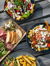 A high-quality, overhead flat-lay shot of a pub-style sharing feast on an outdoor wooden table. The image features loaded nachos, loaded fries, a charcuterie and bread board, and a side of chips, all captured in natural, flattering daylight.