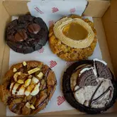 A high-quality, overhead product shot of four distinct, gourmet-style cookies arranged in a branded takeaway box. The cookies feature varied toppings including chocolate chunks, a caramel-like center, and cookie pieces, presented clearly against the branded parchment paper.