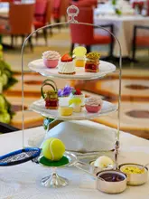 A high-quality, professional promotional photograph of a Wimbledon-themed afternoon tea service at The Peninsula London. The image features a tiered stand with delicate pastries, a chocolate tennis ball, and a mini racket, set against a blurred, elegant hotel interior.