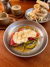 A top-down, slightly cluttered shot of a brunch spread on a wooden table, featuring a hot honey jamon toast as the primary subject, accompanied by a stacked sandwich and ceramic cups of chai latte.