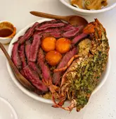 A high-quality, top-down food photography shot featuring a surf and turf rice bowl with sliced steak, a whole lobster tail, and three egg yolks, served in a white bowl on a clean, light-colored surface.