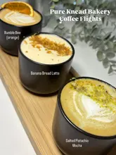 A high-quality, well-lit close-up shot of a coffee flight featuring three distinct lattes and mochas served on a wooden board. The image is clean, bright, and clearly showcases the toppings and latte art of each drink, making it an effective promotional image for a cafe.