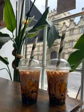 A close-up, eye-level shot of two bubble tea drinks sitting on a wooden table in front of a window, with a blurred view of a historic building in the background.