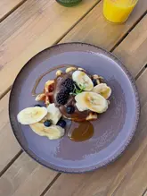 A top-down, overhead shot of a waffle topped with Nutella, banana slices, blackberries, and salted caramel sauce, served on a wooden table with a glass of orange juice visible in the corner.