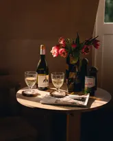 A beautifully composed, atmospheric lifestyle shot featuring a table set for two with white wine and a tin of olives, bathed in warm, golden-hour light. The image evokes a sense of relaxation and luxury, typical of a high-end hotel or aparthotel stay.