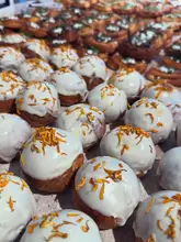 A high-quality, close-up shot of a bakery display case filled with numerous pastries topped with white icing and orange petals. The image uses a shallow depth of field to focus on the front items while creating a sense of abundance in the background.