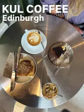 A clean, overhead flat-lay photograph taken at a coffee shop, showcasing a latte with latte art, an iced coffee, a pastry, and a slice of cheesecake on a brushed metal table.