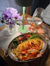 A top-down shot of a lobster linguine dish served in a metal pan, accompanied by a glass of white wine and a vase of flowers on a marble table, presented as part of a listicle about London pasta spots.