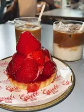 A close-up shot of a strawberry tart served on branded paper, accompanied by two iced coffees in a cafe setting.