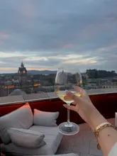 A high-quality, aspirational shot taken from a rooftop terrace in Edinburgh. The image features a hand holding a glass of white wine against a backdrop of the city skyline at sunset, effectively capturing the atmosphere and the venue's primary selling point: the view.