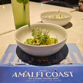 A high-quality, well-lit shot of a ravioli dish from a tasting menu, presented directly on the menu card, accompanied by a cocktail.