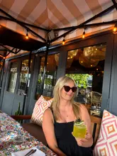 A photo of a woman enjoying a cocktail at an outdoor table at Rick's. The setting is a casual, stylish terrace with a patterned tablecloth and a view of the venue's entrance.