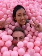 A bright, high-quality selfie of two people submerged in a pink ball pit at an immersive exhibition venue. The image effectively captures the fun, playful atmosphere of the experience.