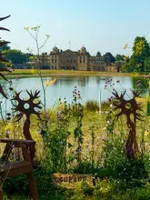 A beautifully composed, scenic shot taken at the RHS Badminton Flower Show, featuring a grand country house in the distance viewed across a lake. The image uses foreground wildflowers and artistic metal sculptures to frame the subject, creating a sense of depth and atmosphere.