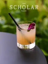 A professionally captured, high-quality still image of a cocktail garnished with a flower, set against a soft-focus garden background. The image effectively promotes the venue's summer atmosphere.
