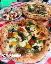 A top-down, brightly lit shot of two pizzas and a side salad served on a classic red and white checkered tablecloth, promoting the new Pizza Pilgrims branch in Bristol.