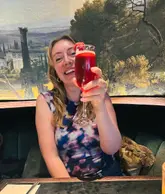 A portrait of a diner holding a cocktail glass in front of their face, set against a decorative mural in a restaurant.