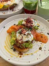A top-down shot of an avocado toast dish topped with poached eggs, smoked salmon, and microgreens, served at a cafe. The table also features other drinks and plates in the background.