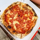 A high-angle, close-up shot of a pepperoni pizza served in a cardboard takeaway box, resting on a wooden table. The pizza features a blistered, charred crust and a generous amount of melted cheese and pepperoni slices.