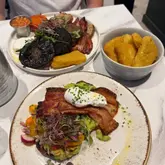 A top-down shot of a brunch spread featuring a full breakfast plate with black pudding, mushrooms, and bacon, alongside a plate of avocado toast topped with a poached egg and a side bowl of hash browns, set on a white marble table.